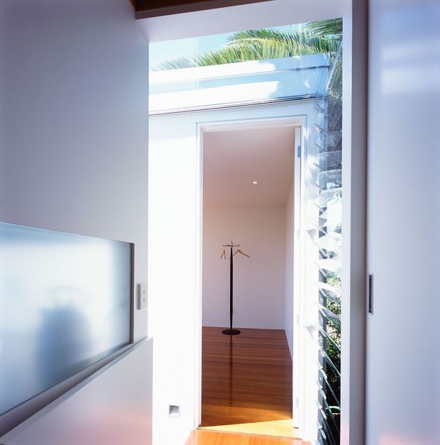 Bright contemporary interior view toward an open doorway leading to a small room or passage with warm timber flooring and a minimalist coat stand. A large frosted or reflective glazed panel is visible on the left, while vertical louvres and tropical foliage appear beside the doorway on the right. White walls, strong daylight, and a timber ceiling detail create a clean, airy transition space.