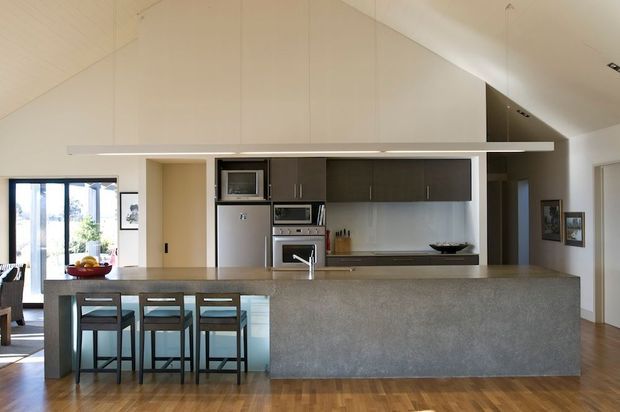 A contemporary open-plan kitchen dominated by a large grey stone island with seating for three. The room has dark fixed cabinetry, integrated stainless-steel appliances, a minimalist splashback, warm timber flooring, and a high sloping ceiling. Dining and living areas are visible beyond the kitchen.