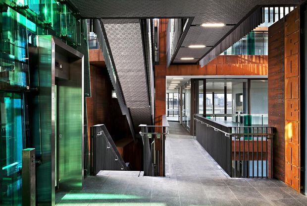 Interior view of a contemporary multi-level commercial or institutional building, featuring a broad circulation corridor and stairwell. The space combines dark tiled flooring, exposed structural and services elements, black metal balustrades, timber-toned wall cladding, extensive glazing, and dramatic green-tinted glass or lighting at the stair and lift zone.