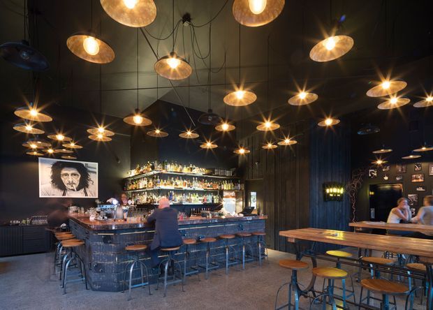 A moody, industrial-style bar and dining venue with a long central bar, extensive back-bar bottle display, high dark ceiling, exposed pendant wiring, and numerous warm metallic pendant lights. Timber communal tables and stools occupy the foreground, while framed artwork and lounge seating appear around the perimeter.