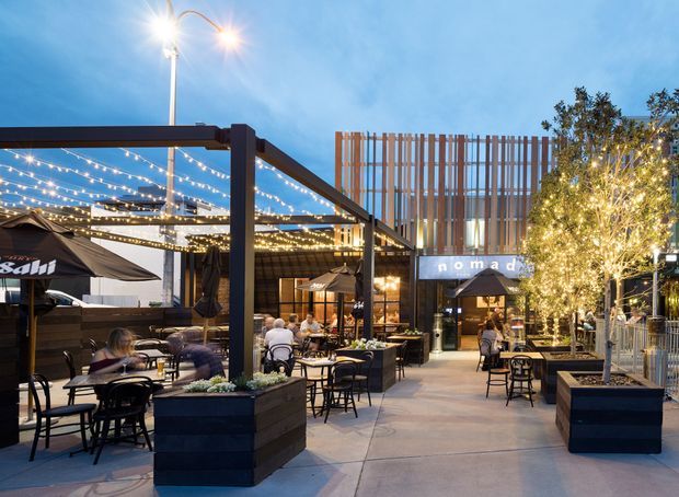 Dusk view of an outdoor commercial dining precinct with multiple café and restaurant seating areas, black steel pergola structures, overhead festoon lighting, large dark planters with small trees, umbrellas, and a prominent Nomad venue sign. A vertical timber-look screen forms part of the backdrop, with illuminated interiors visible beyond the pedestrian circulation space.
