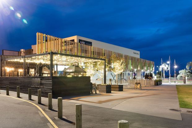 Night-time exterior view of a contemporary waterfront commercial and hospitality precinct. The image shows a low-rise modern building with illuminated timber-like vertical screening, extensive glazing, a covered outdoor dining terrace with festoon lighting, paved pedestrian areas, bollards, landscaping, and marina-style lighting in the background.