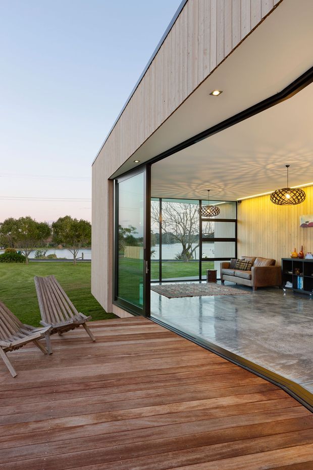Contemporary single-storey home opening onto a timber deck through expansive black-framed sliding glass doors. The interior visible beyond is a polished concrete living room with timber wall panelling, leather seating, patterned pendant lights, and views toward a landscaped lawn and lake.