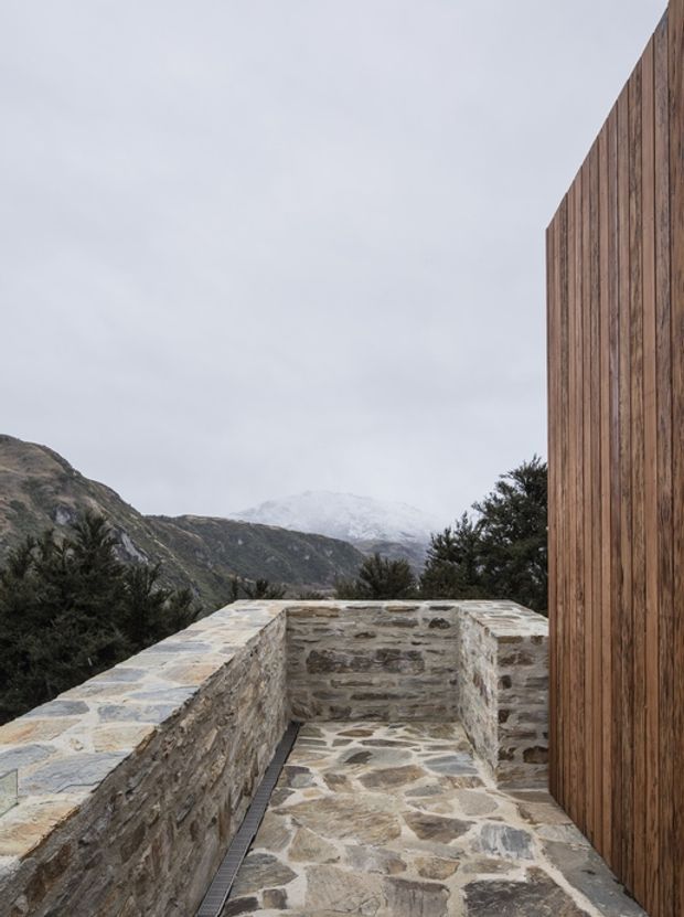 Exterior architectural view of a narrow stone-paved terrace or walkway enclosed by low, rough-stone masonry walls, beside a tall vertical timber-clad wall. Mountainous terrain and an overcast sky are visible beyond, giving the scene a rugged alpine character.