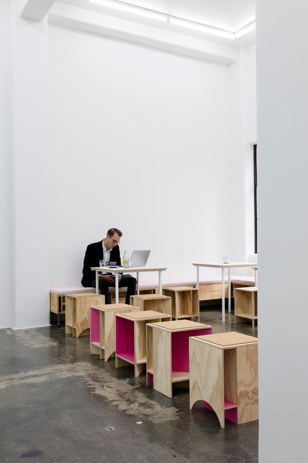A bright, minimalist commercial-style interior with tall white walls, exposed ceiling beams, and linear recessed lighting. The polished concrete floor contrasts with custom pale plywood tables, benches, and sculptural stools featuring vivid pink inset panels. A person works at a laptop in the background, suggesting a flexible studio, workspace, or showroom environment.
