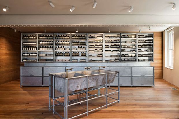 A brightly lit retail showroom with a long wall of neatly arranged product shelving, metal drawer cabinetry below, and a central industrial-style display island with multiple basins or compartments. The space features warm timber flooring and wall panelling, exposed metal fixtures, neutral cabinetry, track lighting, and a large window on the right.