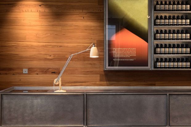 A warmly lit contemporary commercial or retail interior vignette featuring a full-height timber wall with horizontal boards, a slim task lamp on a dark counter, framed abstract artwork, and extensive built-in shelving displaying rows of identical bottles or containers. The foreground includes dark grey textured fixed cabinetry or counter fronts with metal edging. The composition is compact, refined, and strongly material-focused.