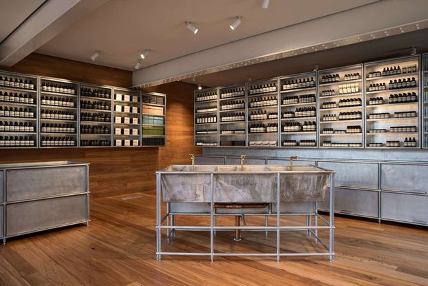 A spacious retail-style interior with extensive wall-mounted shelving displaying rows of small bottles, a central metal island display or counter, and large metallic base cabinets. The room combines warm wood flooring and wall panelling with brushed metal fixtures and cabinetry, illuminated by ceiling track lights.