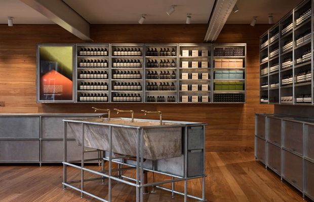A warmly lit, industrial-style retail or tasting space with extensive wall-mounted shelving displaying rows of bottles and boxed goods. The room has rich dark timber wall cladding and timber flooring, exposed metal-framed cabinetry and counters, a central metal island with multiple brass taps, and track lighting. A framed artwork is mounted on the left wall.