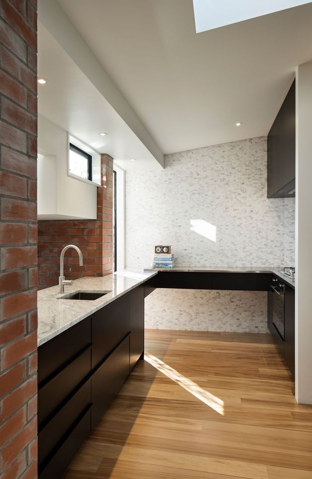 A bright, narrow contemporary kitchen with a U-shaped dark joinery layout, pale stone benchtops, and a stainless-steel gooseneck sink tap. The walls combine exposed red brick with a light small-format mosaic tile splashback, while warm timber flooring runs throughout. Recessed ceiling lights and high windows bring in natural light, with a small skylight or ceiling opening visible above.