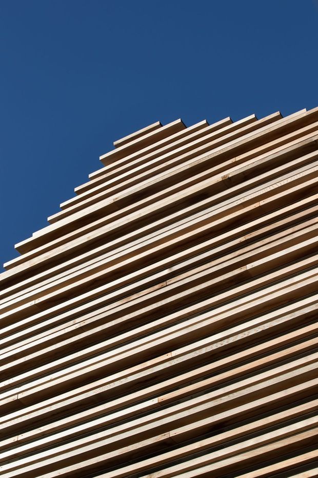 Close-up upward view of a contemporary building facade clad in closely spaced horizontal timber slats, creating a strong layered linear pattern against a clear deep-blue sky. The image is dominated by the exterior wall treatment and its warm natural wood tones.