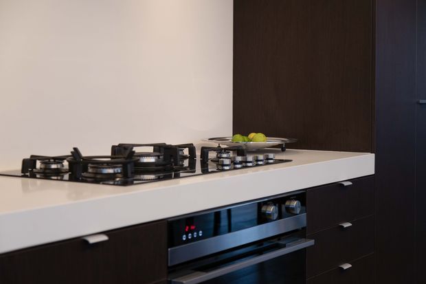 Close-up view of a contemporary kitchen island with a pale stone benchtop, built-in gas cooktops, and an integrated stainless-steel oven. Dark timber-look cabinetry and a dark vertical feature panel form the backdrop, creating a high-contrast, streamlined fit-out.