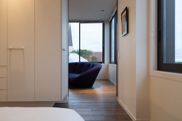 A narrow, softly lit interior passage viewed from a bedroom, with a large built-in light timber wardrobe on the left and a partial bed in the foreground. The corridor leads to a small sitting area with a dark upholstered lounge chair beside full-height windows. Warm timber flooring, pale walls, white trim, framed artwork, and dark window frames create a calm contemporary interior.