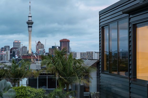 View from a rooftop terrace or upper-level outdoor area toward a dense city skyline, with Auckland's Sky Tower prominently visible in the distance. The foreground includes tropical palm planting, a glass balustrade, and a dark contemporary building facade with large windows and horizontal cladding at right. The scene is photographed under overcast blue-grey skies with warm interior light visible through the glazing.