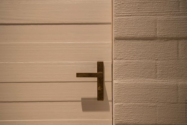 Close-up architectural detail of a minimalist light timber-toned door with horizontal recessed panel lines and a dark bronze or black metal lever handle. The door meets a textured beige plaster or stucco wall, with warm directional lighting emphasizing the surface textures and handle shadow.