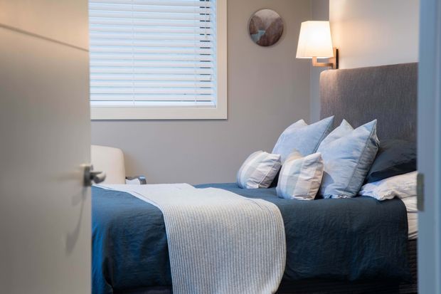 A compact, calm bedroom viewed through a partially open door. The room features a neatly made bed with a tall upholstered grey headboard, dark blue bedding, pale blue and white layered pillows, and a light textured throw. A white roller-blind window provides soft daylight, while a wall-mounted lamp adds warm illumination. Neutral grey walls and minimal artwork complete the understated interior.