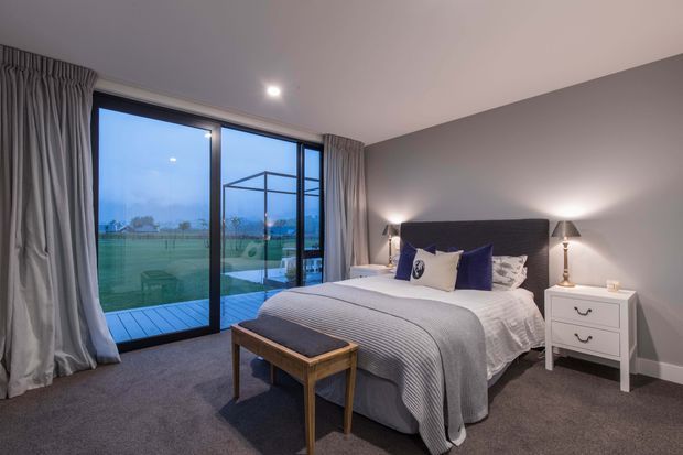 A contemporary bedroom with a neatly made bed, dark upholstered headboard, bedside tables and lamps, and a bench at the foot of the bed. Floor-to-ceiling sliding glass doors open to a small deck and a green outdoor area, with full-length grey curtains framing the glazing. The room has soft grey carpeting, light neutral walls, and blue and white bedding accents, creating a calm, refined atmosphere.