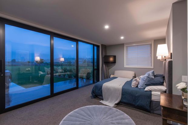 A modern, compact bedroom with a low-profile upholstered bed, layered blue and grey bedding, bedside table and lamp, wall-mounted television, and grey carpet. Full-height dark-framed glazing opens the room visually toward an outdoor pool or terrace, while a roller blind covers the smaller window. Recessed ceiling lights and a neutral palette create a calm, contemporary atmosphere.