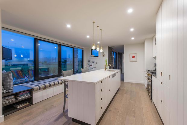 Contemporary open-plan kitchen with a large white island, integrated drawers, extensive white cabinetry, light timber-look flooring, and a built-in window seat beside full-height glazing. Pendant lights hang above the island, while the glazing offers an expansive blue-toned exterior view. The space is clean, bright, and refined with a neutral palette and strong indoor-outdoor connection.