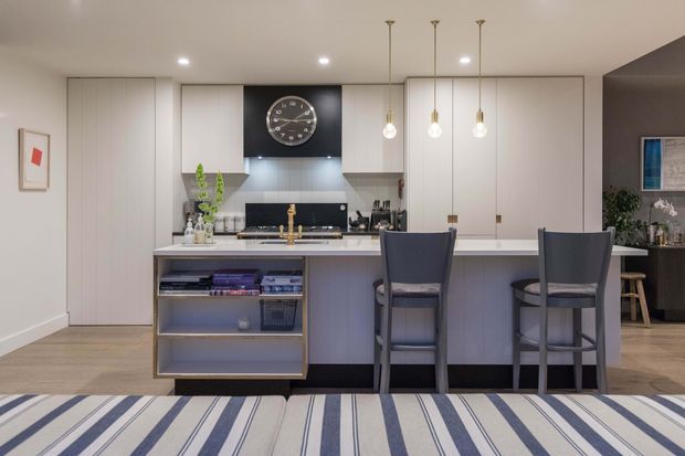 A contemporary open-plan kitchen and dining area viewed from the living space. The room features a large pale kitchen island with integrated open shelving, white flat-panel cabinetry, a dark feature backsplash and clock, pendant lighting, timber-look flooring, and two dark upholstered bar stools. A striped rug occupies the foreground, while a darker adjoining living or dining area is visible at right.