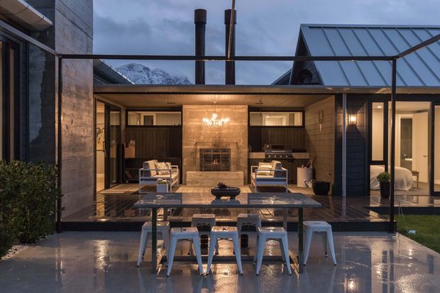 A contemporary residential courtyard viewed from a wet outdoor terrace at dusk. The open-sided living area features a central stone fireplace, built-in timber cabinetry, lounge seating, warm pendant lighting, and extensive dark-framed glazing. A dining table sits in the foreground, while pitched metal roof forms and a mountain backdrop are visible beyond.