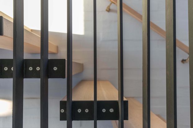 Close-up architectural detail of a black metal balustrade or stair guard viewed through vertical bars. A timber stair and matching timber handrail are visible behind the railing, with pale walls and strong natural light creating a clean, modern interior composition.