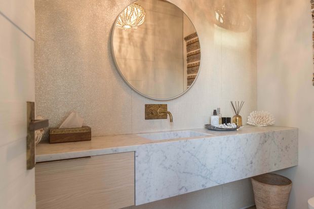 A refined bathroom vanity area with a floating pale timber vanity and a thick white marble benchtop featuring grey veining. A large round mirror, wall-mounted brass tapware, textured light wall finish, decorative lighting reflected in the mirror, and woven accessories create a warm, luxurious interior.