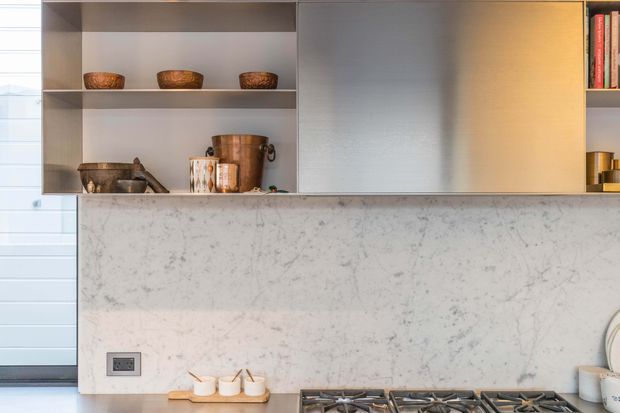 Close-up view of a refined contemporary kitchen featuring a pale veined stone splashback and benchtop, integrated grey cabinetry, open display shelving with ceramic and timber vessels, warm under-cabinet lighting, and a built-in gas cooktop in the foreground. A glazed or louvred opening is visible at the left edge.