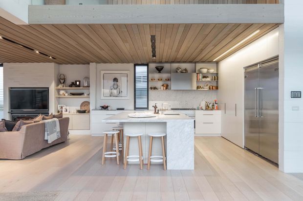 Open-plan contemporary kitchen and living area with a central marble-look island and seating for four, white cabinetry, integrated appliances, pale timber flooring, and a warm timber-lined ceiling beneath a mezzanine or upper-level void. A living room with sofa and television is visible to the left, while open shelving and a textured light backsplash add detail to the kitchen.