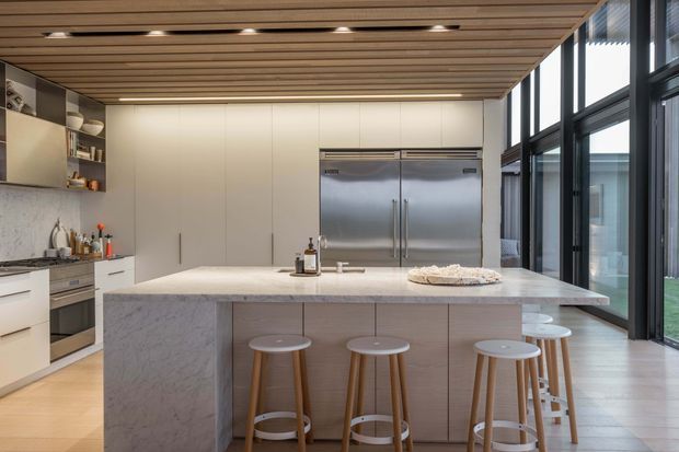 A bright, spacious contemporary kitchen centered on a large marble-look island with seating for five or more. The kitchen features pale fixed cabinetry, integrated appliances, a stainless-steel double-door refrigerator, expansive black-framed glazing, and a timber slatted ceiling with recessed lighting. The palette is light and neutral with warm timber accents.