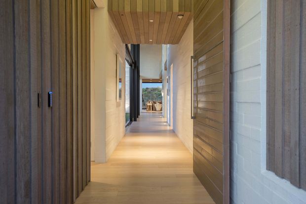 A long, narrow interior hallway with a warm contemporary design. The corridor features pale timber flooring, white rendered or painted walls, extensive vertical and horizontal timber wall cladding, and a timber-lined ceiling with recessed downlights. Dark-framed glazing and doorways run along the hall, leading toward a bright opening and an outdoor view at the far end.