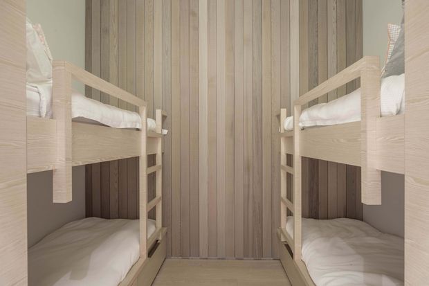 Compact bunk-bed room with two sets of built-in timber bunk beds facing each other. The walls are clad in vertical natural timber boards, with pale timber bed frames, integrated ladders, white bedding, and a simple timber floor. The symmetrical layout and enclosed scale suggest a practical children’s or shared sleeping space.