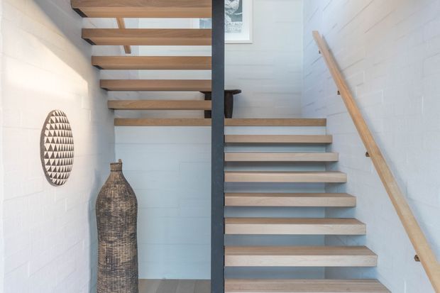 A bright, minimalist stairway with open-riser timber treads supported by a dark central steel spine. White painted masonry walls, a timber handrail, and a small skylight create an airy, contemporary interior. Decorative woven objects sit along the left wall.