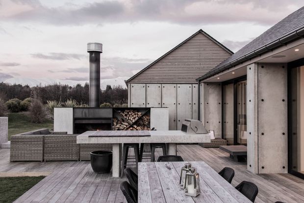 A sophisticated outdoor entertaining area with a timber deck, long dining table, built-in barbecue and fireplace zone, outdoor kitchen cabinetry, and adjoining modern house forms. The design uses weathered timber, pale concrete or stone surfaces, dark metal detailing, and a pitched roof, set against landscaped grounds under an overcast sky.