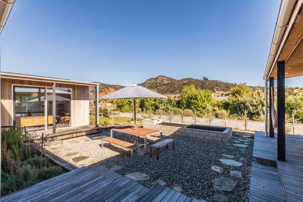 A sunny landscaped courtyard and outdoor entertaining area between modern timber-clad structures. Timber decking, gravel paving, stepping stones, raised concrete-block garden beds, outdoor dining furniture, and a large umbrella frame views toward a dry, rocky hillside. The architecture features simple low-pitched roof forms, extensive glazing, timber soffits, and dark slender posts.