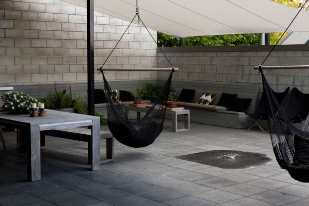A contemporary covered courtyard or patio with large grey tiled paving, concrete block privacy walls, a stretched shade canopy, black hanging hammocks, and built-in bench seating with cushions. A substantial outdoor dining table occupies the foreground, while potted plants and a small wet patch add lived-in detail. The space has a sheltered, relaxed outdoor entertaining atmosphere.