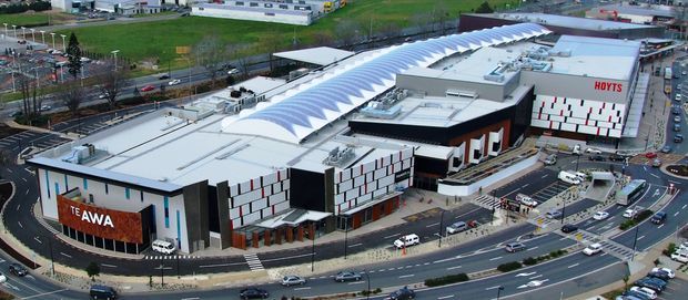 Aerial view of a large contemporary commercial entertainment complex in an urban setting. The complex features expansive flat-roofed volumes, a prominent long ribbed or arched translucent roof canopy, patterned white and dark facade panels, and visible signage for Te Awa and HOYTS. Multi-lane roads, parking areas, and neighboring commercial buildings surround the site.
