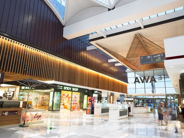 Bright contemporary shopping-centre interior with retail storefronts, polished tiled flooring, a dramatic timber slatted feature wall and balustrade, a suspended sculptural timber ceiling installation, extensive skylights, and open circulation space with wayfinding signage.