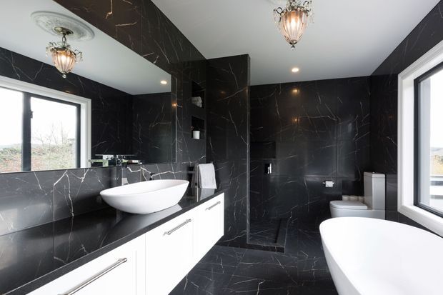 Luxurious contemporary bathroom clad extensively in dramatic black marble-look slab surfaces with contrasting white fixtures and vanity cabinetry. The room includes a double vanity with vessel basin, freestanding bathtub, toilet, large windows, mirrored wall, recessed lighting and decorative pendant fixtures.