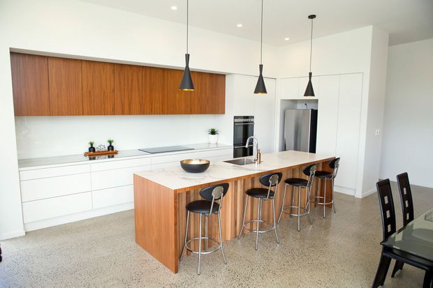 Bright contemporary open-plan kitchen with extensive white handleless cabinetry, a long warm timber feature cabinet, and a matching timber island with seating for four. Three black pendant lights hang above the island, while integrated appliances and a stainless-steel refrigerator line the rear wall. The space opens toward a dining area with a dark dining table and chairs. Polished exposed aggregate or terrazzo-like flooring and white walls create a clean, spacious feel.