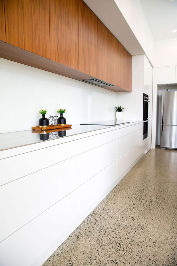 Bright contemporary kitchen with extensive white handleless cabinetry, a long white benchtop, warm vertical timber overhead cabinets, integrated black appliances, and a polished speckled concrete or terrazzo-like floor. The linear composition and minimal styling create a clean, modern interior.