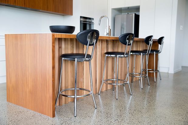 Bright contemporary kitchen with a timber island and matching timber cabinetry, a white benchtop, stainless steel appliances, and a row of black bar stools. The space has polished exposed aggregate or terrazzo-like flooring, white walls, and a clean open-plan layout.