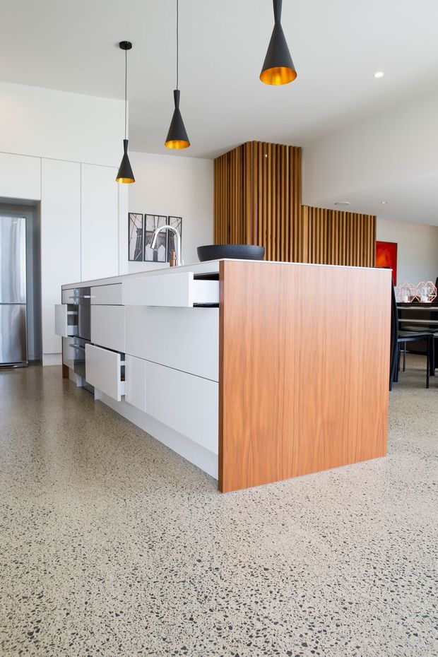 Bright contemporary kitchen with a large white island and warm timber-clad end panel, integrated cabinetry, a stainless-steel tap, and three black pendant lights. A vertical timber slat feature wall extends toward the adjacent dining area. The room has a polished speckled concrete or terrazzo-style floor and a clean, open-plan layout.
