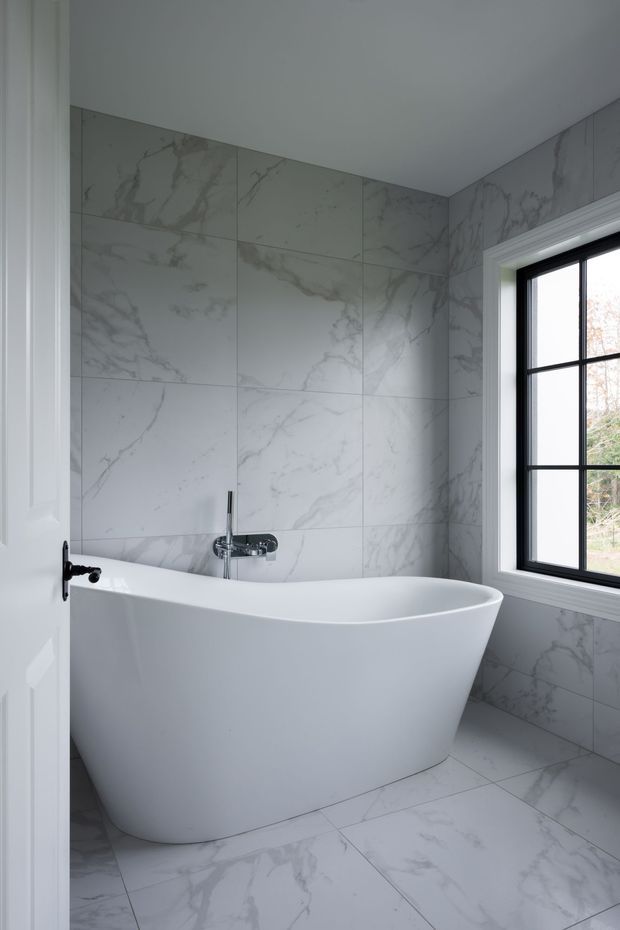Bright, refined bathroom featuring a freestanding white oval bathtub, chrome wall-mounted tapware, and large white marble-look tiles with grey veining across the walls and floor. A black-framed divided-light window provides natural light, while a white paneled door is visible at the left.
