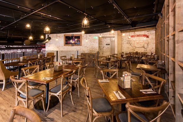 A warmly lit restaurant dining area with numerous timber tables and cross-back wooden chairs, exposed dark ceiling services, pendant cage lights, timber wall cladding, and an exposed brick feature wall. Large vintage map artwork and casual hospitality furnishings create a rustic industrial atmosphere.