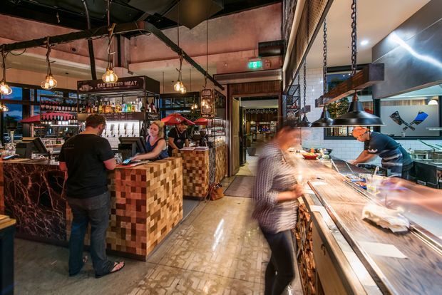 A lively industrial-style restaurant interior showing a bar and customer service counter on the left, an open commercial kitchen and long preparation counter on the right, and a central circulation aisle. The space features exposed dark services and ductwork, pendant lights, black metal shelving and framing, patterned floor tiles, timber mosaic counter cladding, and warm atmospheric lighting. Staff and patrons are present, with motion blur emphasizing the busy hospitality setting.