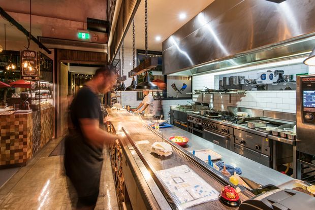 A busy commercial open kitchen viewed from the service counter, with stainless-steel cooking equipment, tiled splashback walls, a large metal extraction canopy, hanging pendant lights, timber and mosaic finishes, and staff working behind the counter. The space has a warm industrial restaurant atmosphere.