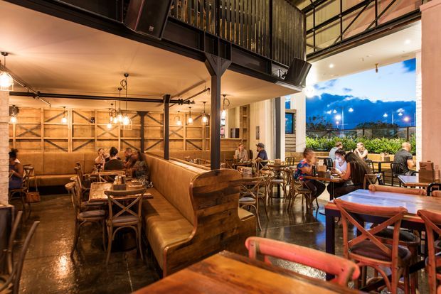 A warmly lit restaurant dining area with exposed black steel framing, timber wall panels, leather banquette seating, rustic timber tables and chairs, and pendant lighting. The open frontage leads to an outdoor terrace with evening sky and planted screening visible beyond.