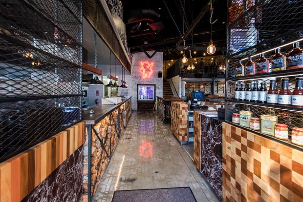 A narrow, atmospheric commercial interior resembling a bar, café, or restaurant, viewed down a central polished concrete aisle. The space features extensive black metal mesh partitions and shelving, dark exposed ceiling services, warm pendant and caged lighting, timber block-patterned counters, dark marble-look panels, displayed bottles and food products, and a glowing red wall sign. The design has an industrial character with warm timber accents and dramatic lighting.