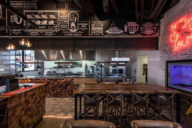A dark, industrial-style restaurant interior focused on an open commercial kitchen and long timber dining counter. The space features exposed black ductwork and services, a chalkboard-style illustrated wall graphic explaining the smoking process, exposed brickwork, white tiled kitchen walls, stainless-steel commercial equipment, timber counter surfaces, stacked firewood, metal-framed stools, pendant lights, and red neon signage.